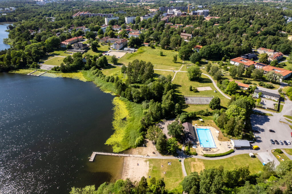 Grönt parkområde vid sjö. I förgrunden brygga och bassäng. 